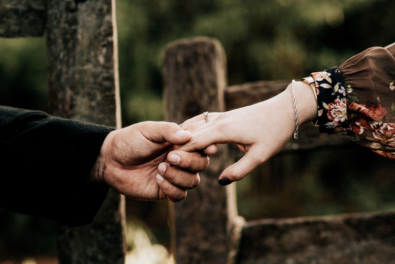 a couple holds hands by a fence, the woman wearing a solitaire engagement ring.