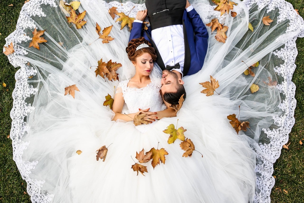 A bride and groom lay down on the grass as leaves fall on them.