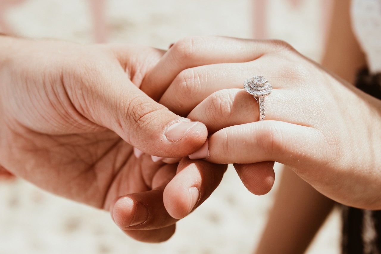 a man holds his fiancee’s hand, inspecting her halo engagement ring. 
