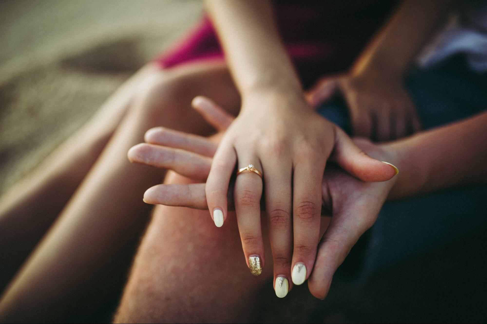 A couple holding hands while sitting outside. The woman wears a gold engagement ring.