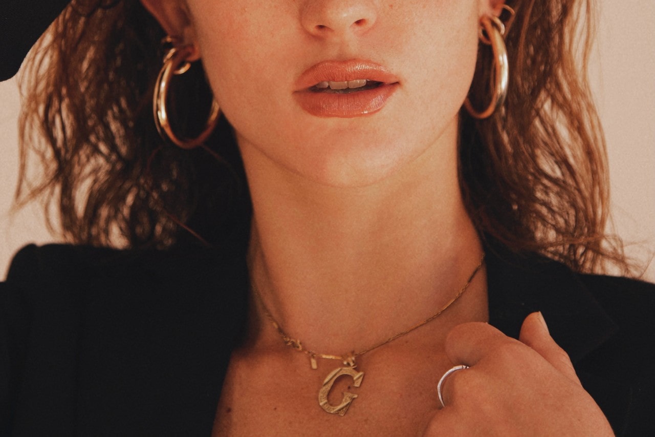 a woman wearing gold earrings, a gold “G” pendant necklace and a gold ring